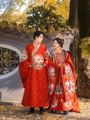 Front three-quarter view of a couple wearing matching Ming Dynasty Chinese wedding Hanfu in scarlet red robes with auspicious embroidery, posing in a traditional Chinese garden with autumn ginkgo leaves