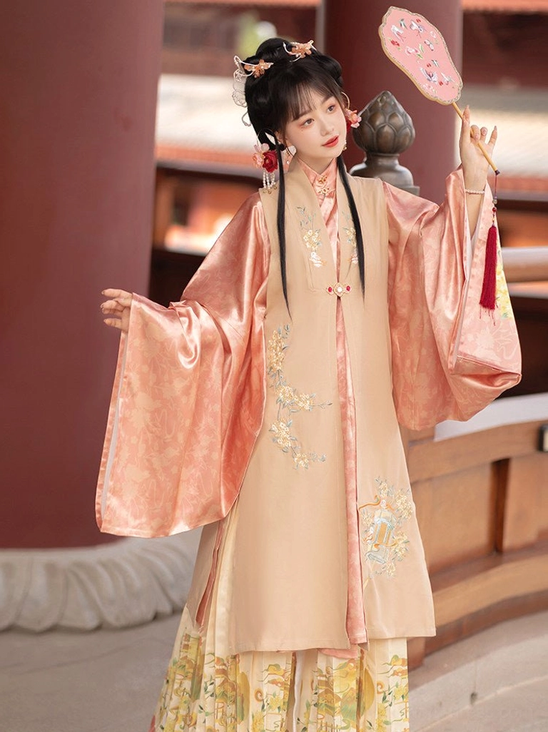 Close three-quarter view of a woman wearing a cream bijia vest and salmon pink Ming-style robe with floral embroidery and coordinated pleated skirt details