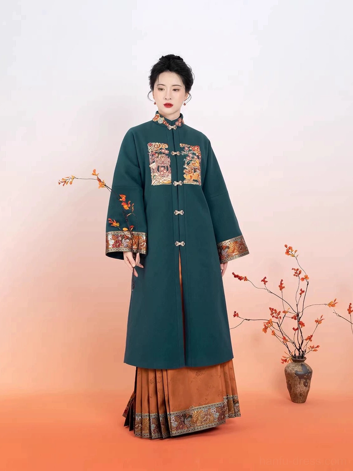 Model wearing a dark teal Ming Dynasty Hanfu robe with embroidered details and a burnt orange pleated mamianqun skirt in a warm studio setting
