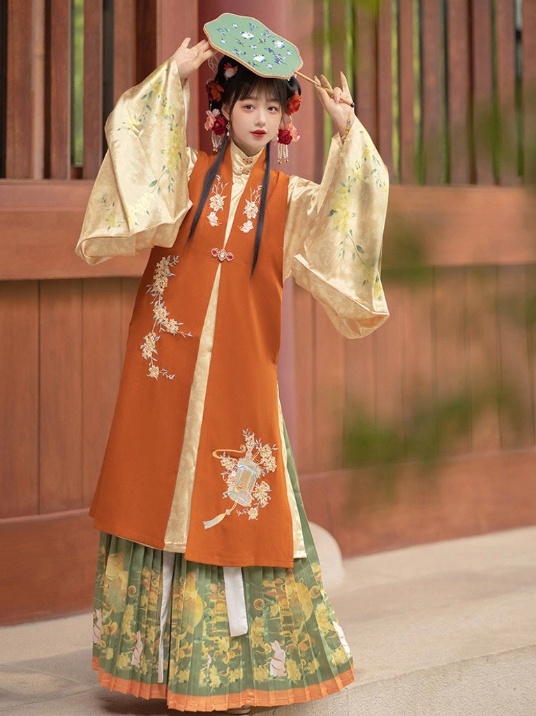 Full-body front view of a woman wearing a three-piece Ming Dynasty hanfu set with terracotta bijia vest, cream robe, green pleated skirt, and embroidered round fan
