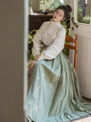 Three-quarter front view of a Ming Dynasty-inspired Hanfu set with an off-white striped stand-collar top, sage green pleated Mamianqun skirt, and bamboo embroidery in a warm traditional interior