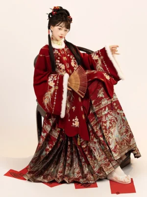 Ming Dynasty inspired red Hanfu Chinese New Year dress shown in a three quarter seated view with white fur effect trim, phoenix embroidery, gold floral details, and a pleated mamian horse face skirt