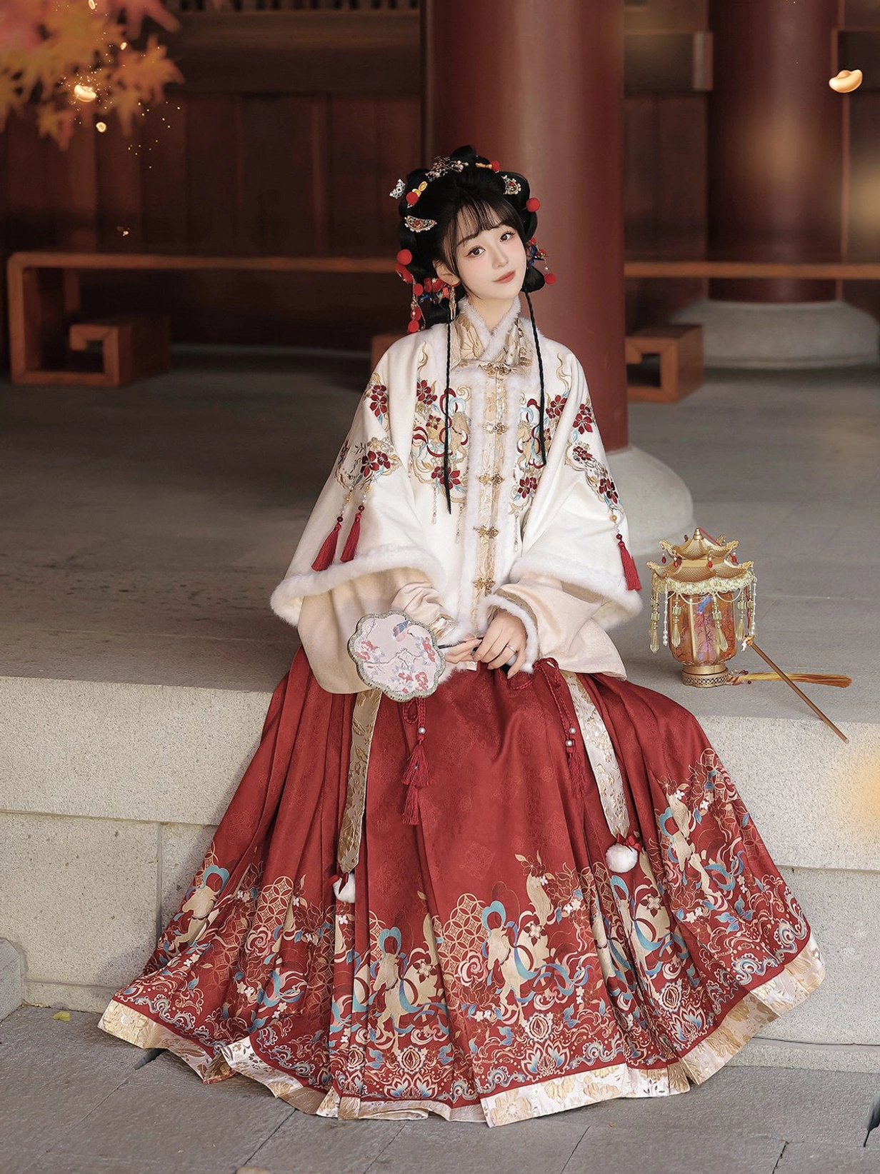 Festive Chinese New Year Hanfu outfit seated in courtyard wearing embroidered pipa sleeve top and red pleated mamian skirt with silk fan