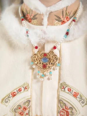 Front close-up of a Ming-style Chinese Hanfu necklace worn on an embroidered fur-trimmed coat, showing the gilded longevity lock pendant, blue center stone, white pearl chain, colorful beads, and dangling pearl tassels