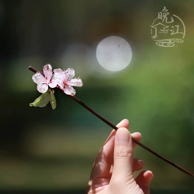 Mini peach blossom Chinese hair accessory held in hand, showing full dark brown hair stick with translucent pink resin flowers and green leaves