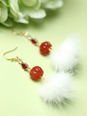 Gold hook mink furball pumpkin bead drop earrings with red gemstone accents displayed beside green hydrangea flowers