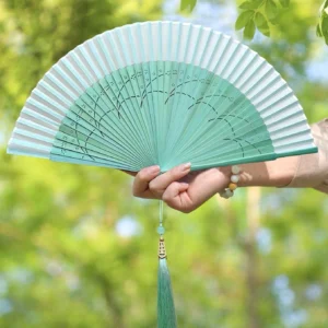 Mint green Chinese folding hand fan with black floral vine pattern, bamboo ribs, jade-like tassel pendant, and outdoor foliage background