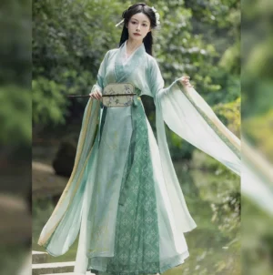 Three-quarter view of a mint green Wei Jin Dynasty Hanfu dress with sheer flowing sleeves, geometric dark green skirt panels, traditional round silk fan, and woodland waterside background