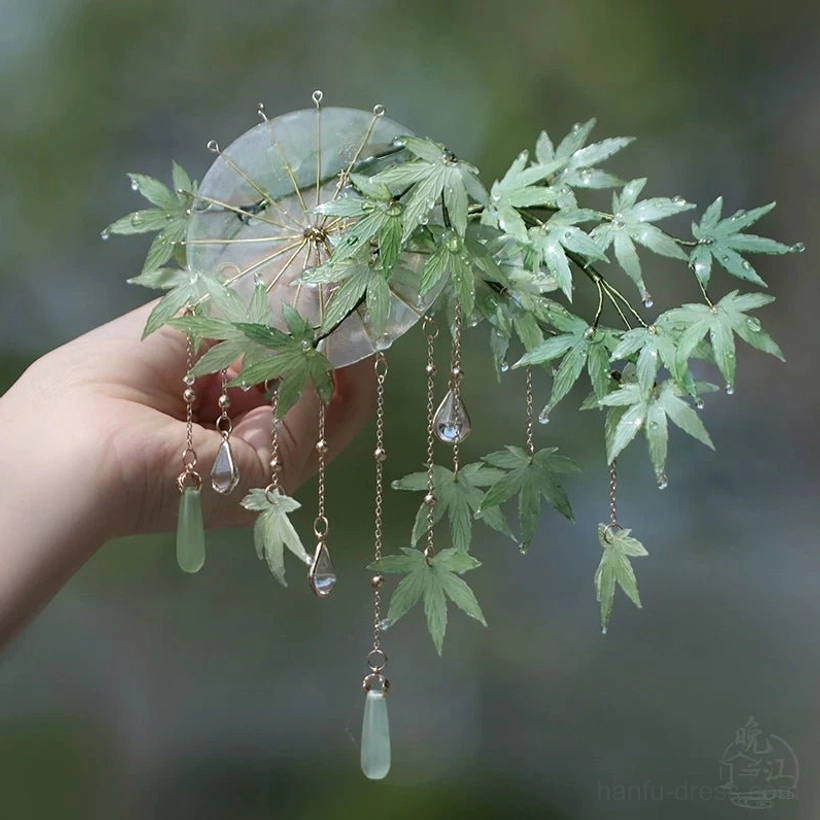 Mint green maple leaf Chinese hair stick with gold tassel chains, teardrop crystal and jade-style pendants held against a blurred green outdoor background