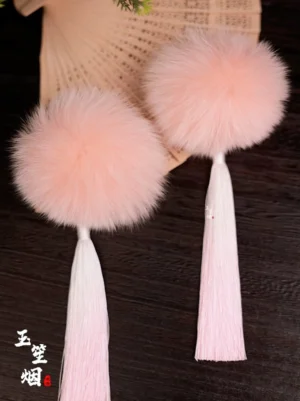 Pair of pink pom-pom tassel Hanfu hair clips displayed on dark wood beside a traditional Chinese folding fan