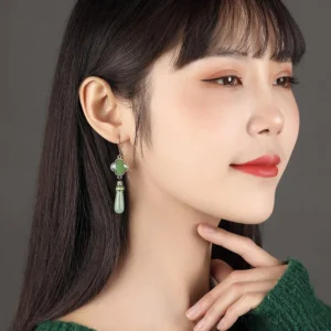 Model wearing light green jade four-leaf clover Chinese earrings with elegant drop pendants and enamel details against a dark green sweater