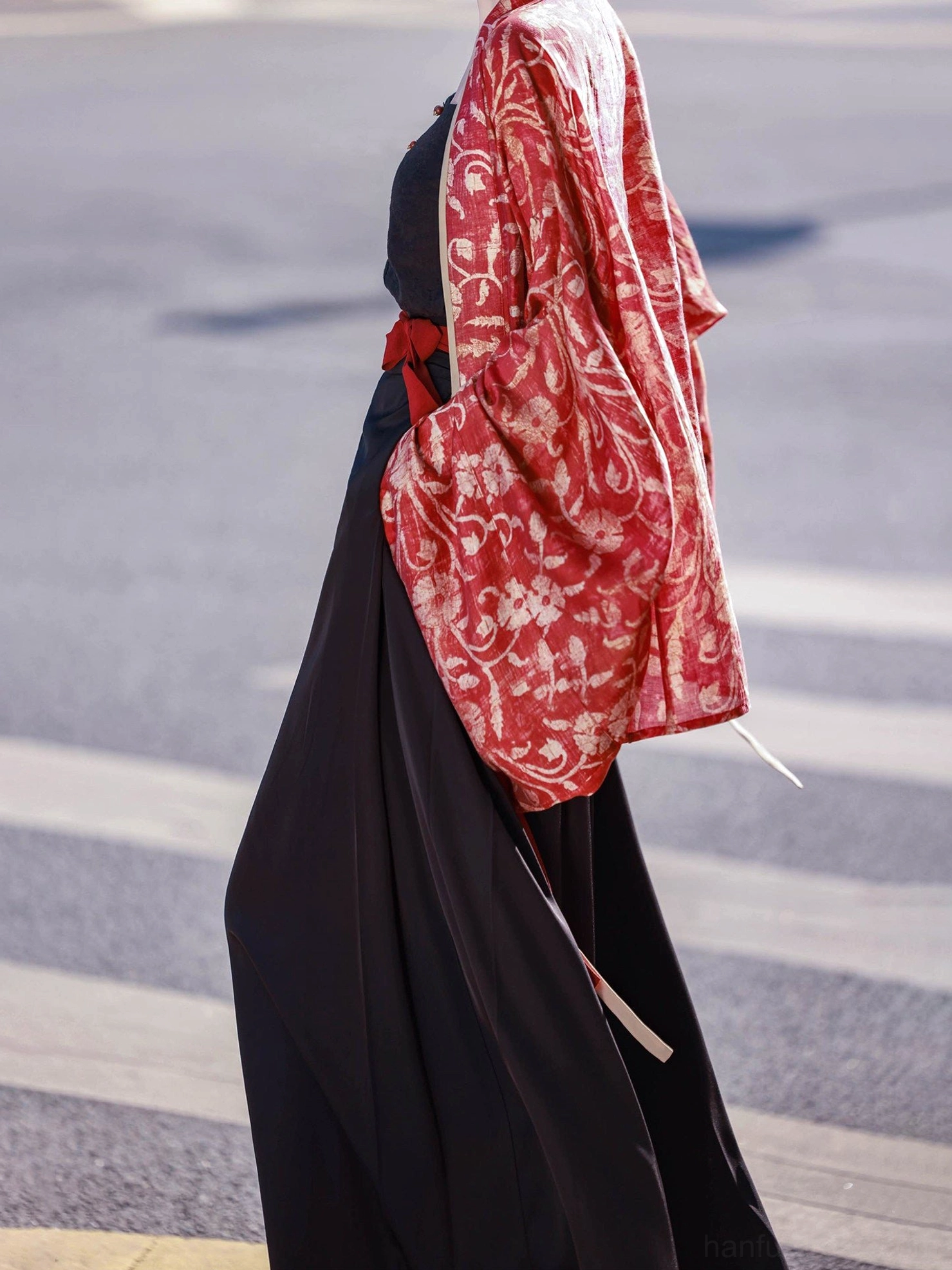 Back three-quarter detail of a red printed Chinese Hanfu cardigan with beige contrast trim worn over high-waisted black Song-style trousers