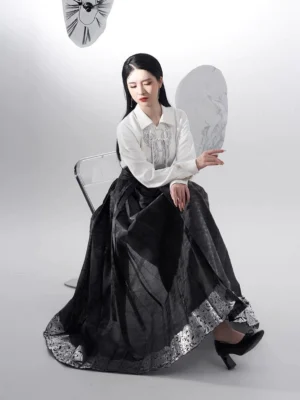 Three-quarter seated view of black modern Chinese hanfu with flowing pleated mamianqun horse face skirt and coordinated silver embroidered hem