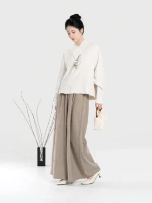 Three-quarter view of a woman modeling a modern Chinese style Hanfu outfit with embroidered shirt, wide-leg pants, and cream pleated handbag