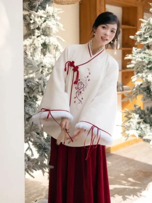 Modern Chinese style Hanfu winter coat shown beside a snowy evergreen tree, featuring a cross-style collar, red trim, floral embroidery, decorative knots, and fur-trimmed wide cuffs