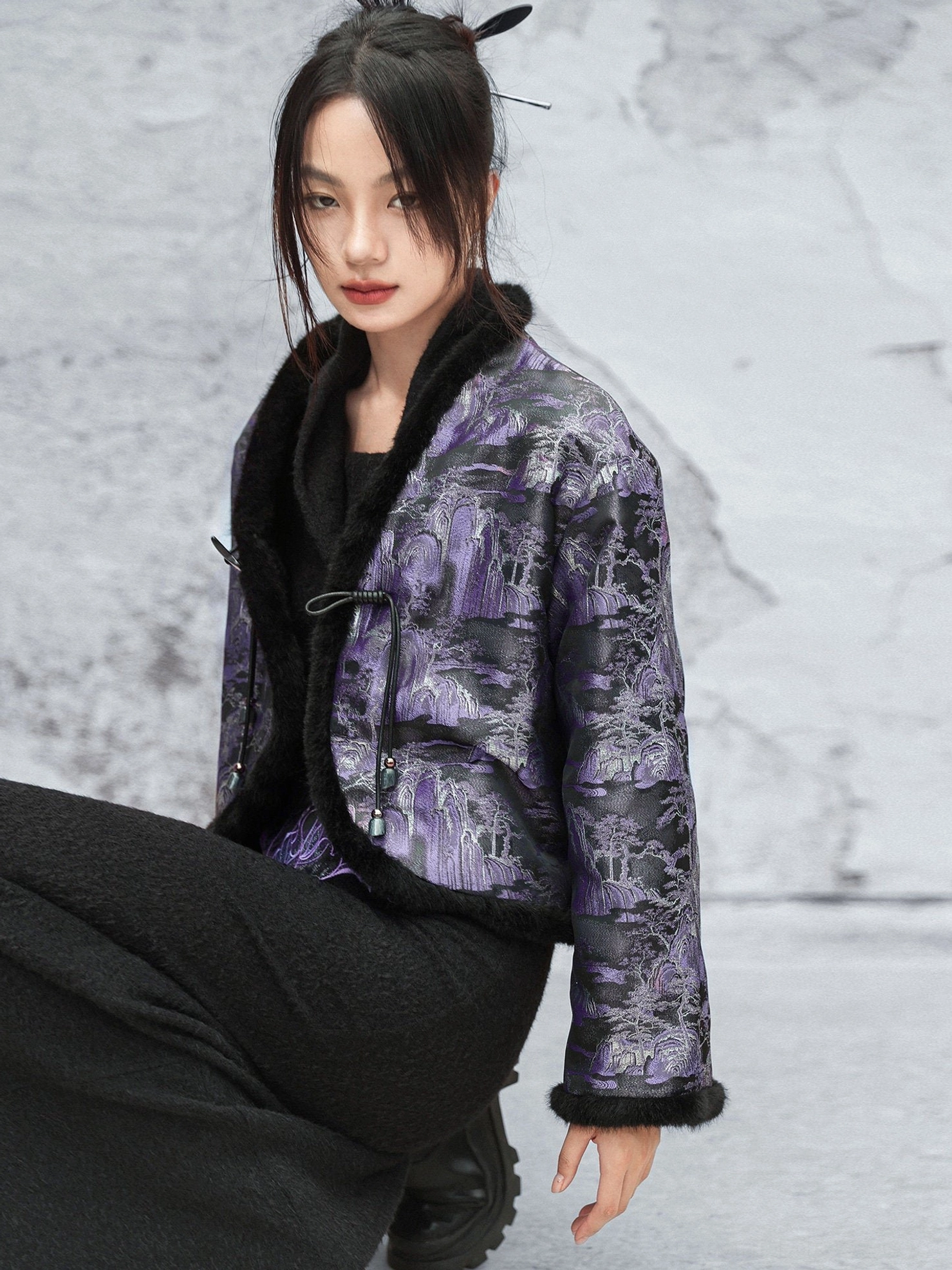 Close-up seated view of a modern Chinese style cropped jacket featuring a dark purple jacquard pattern black fuzzy collar cuffs and traditional frog button closures