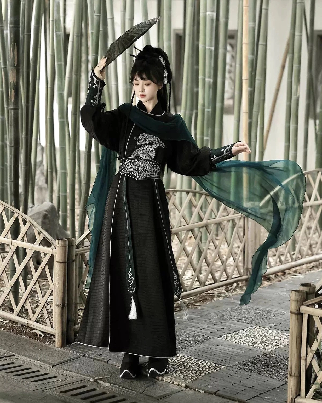 Side rear view of a modern Chinese Hanfu xianxia costume with flowing dark green scarf, silver embroidered waist detail, and black layered robe in a bamboo courtyard