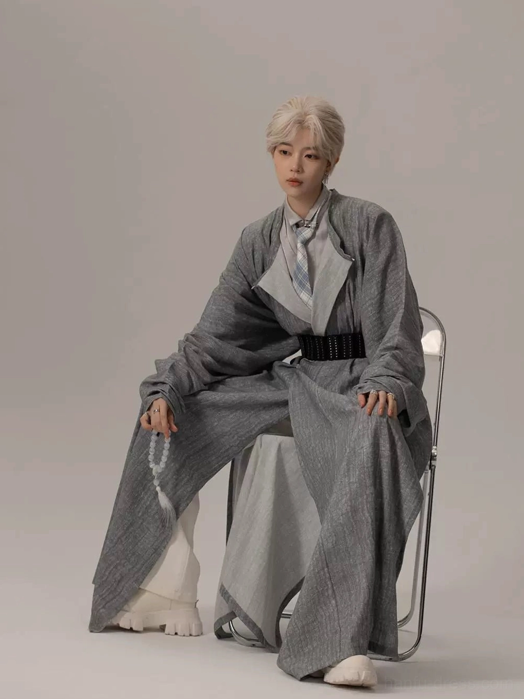 Seated styling view of a modern grey Hanfu robe with oversized sleeves, black waist belt, side opening, wide inner trousers, and decorative tassel accessory