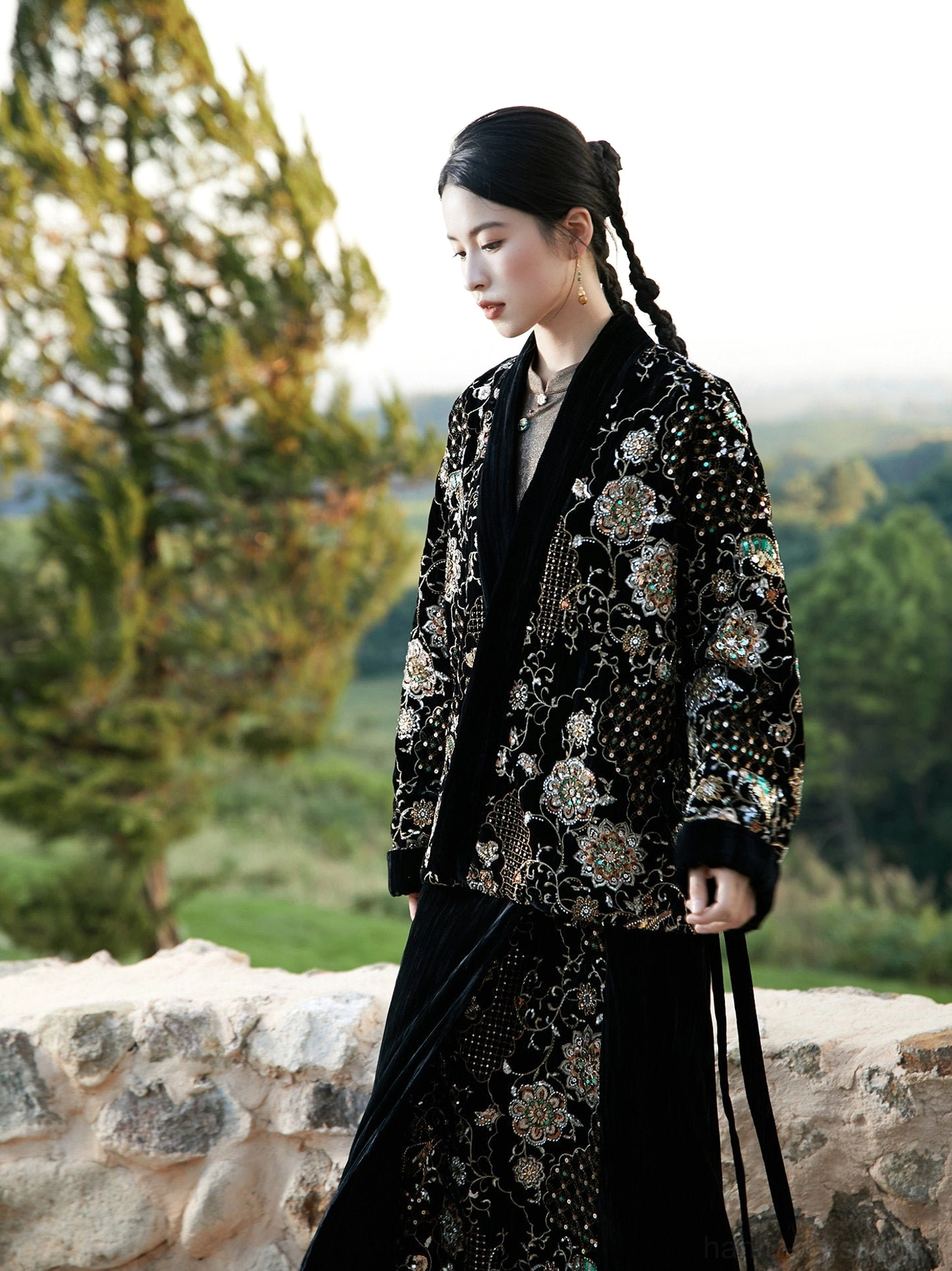 Side view of black modern Hanfu beaded embroidered wool-padded jacket and velvet wrap skirt worn outdoors beside a stone wall and pine tree