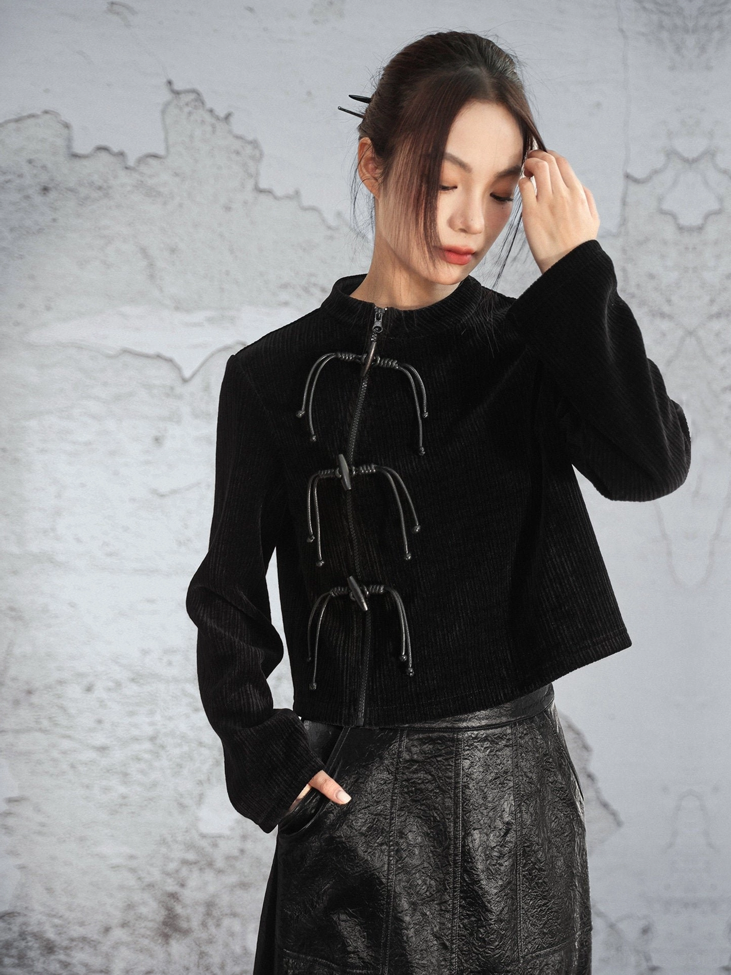 Three-quarter front view of a black modern Hanfu chenille knit sweater with stand collar vertical stripe texture and decorative Chinese frog closures styled with a leather midi skirt