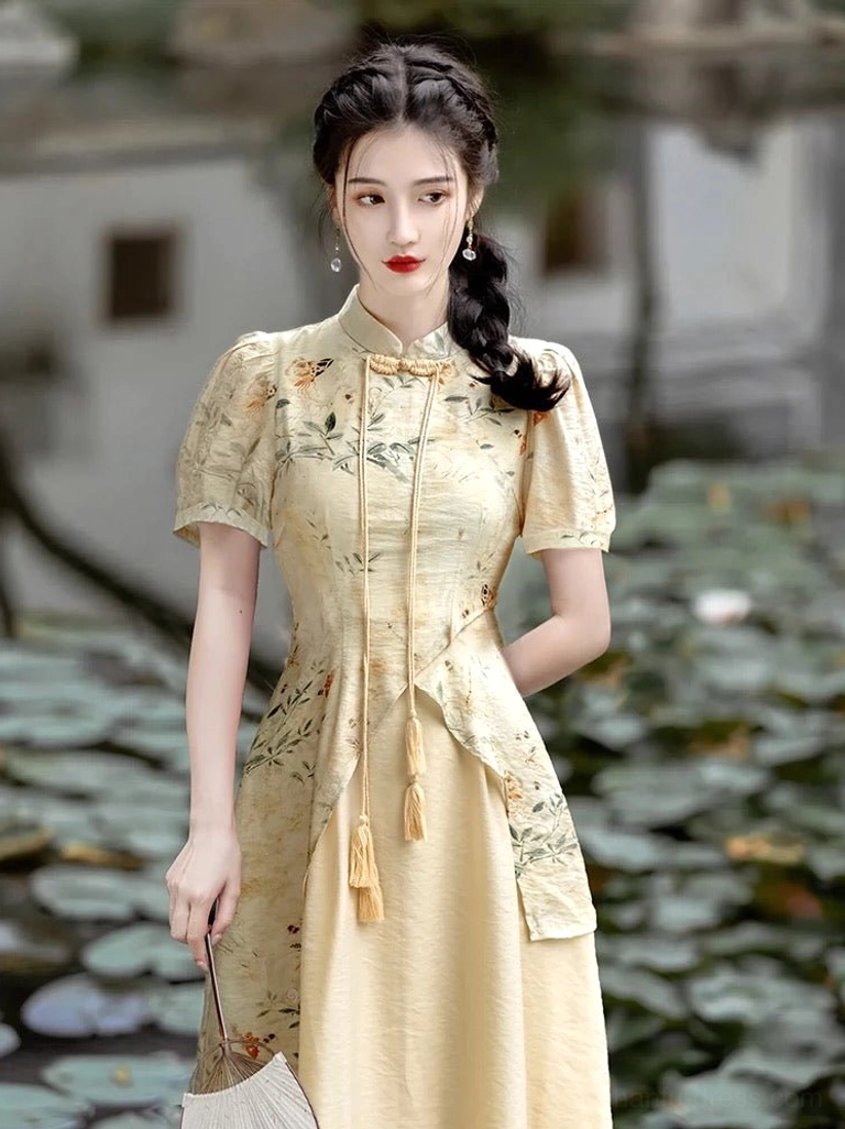 Full front view of a modern Chinese-style Hanfu cheongsam dress with apricot beige fabric, fitted waist, ink-wash floral overlay, stand collar, tassel details, and woven mini handbag