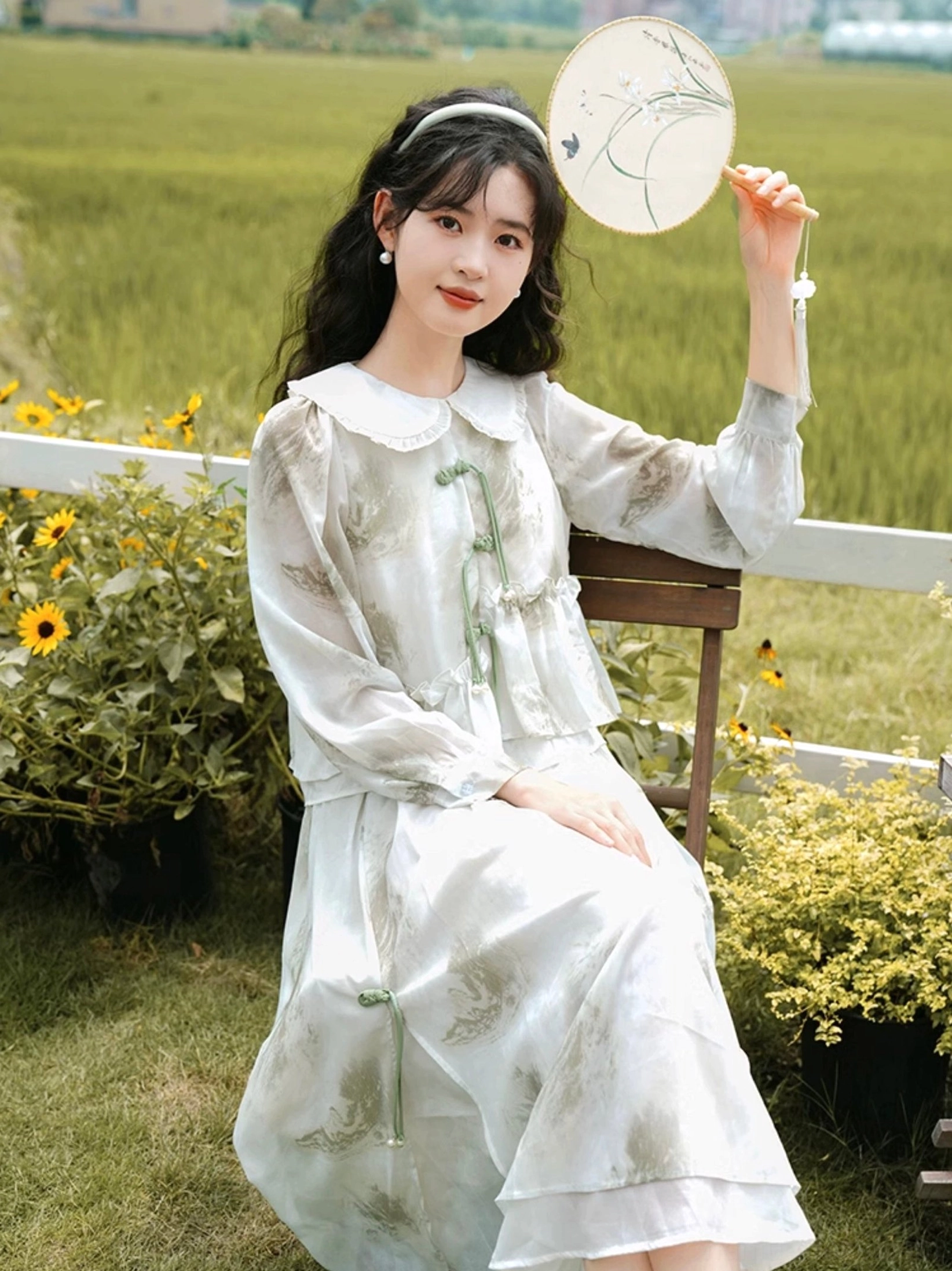 Seated three-quarter view of a Chinese-inspired modern Hanfu dress set with doll collar, frog buttons, sheer sleeves, ink botanical prints, and flowing skirt