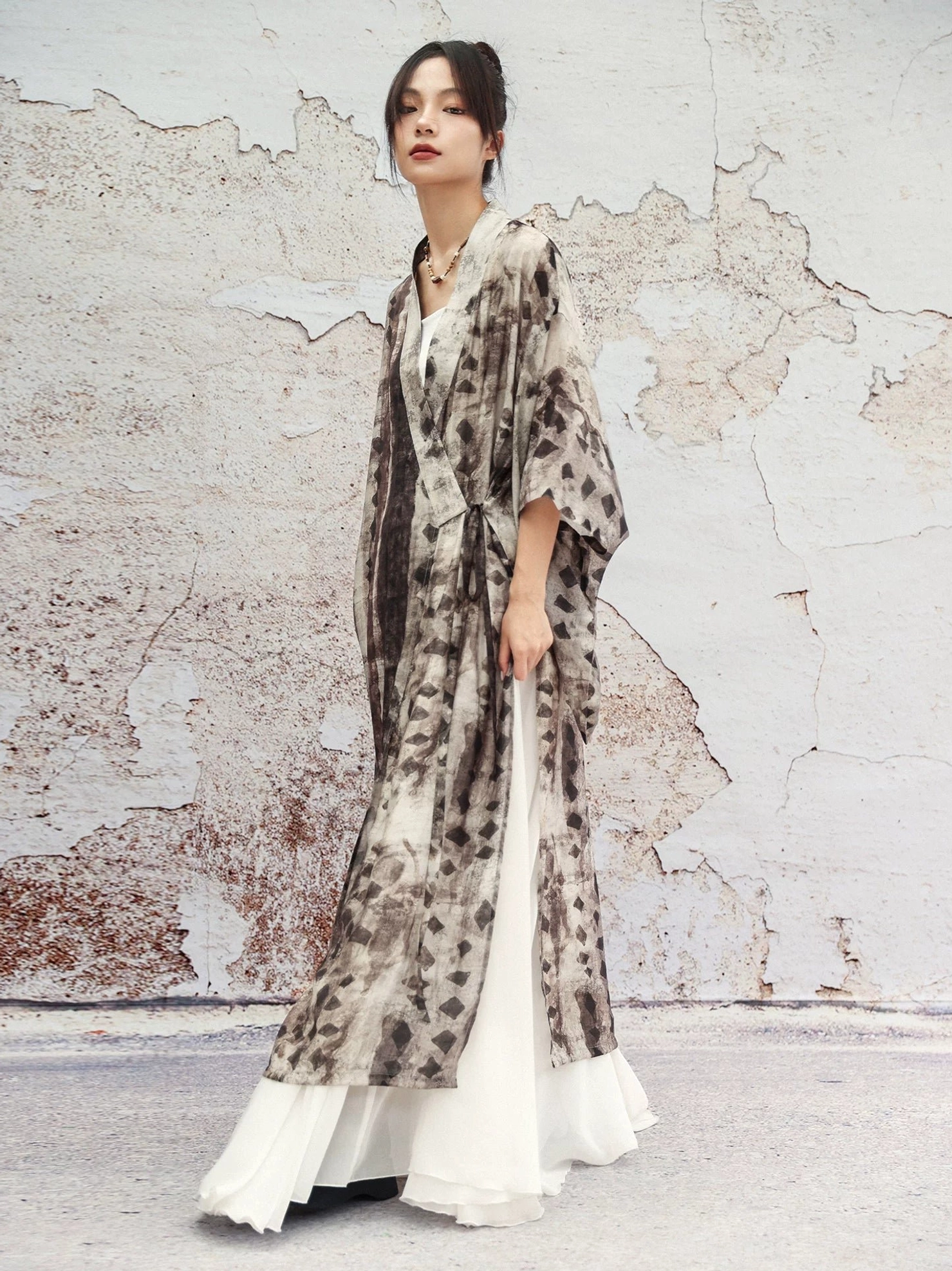 Modern Hanfu long cardigan in earthy geometric ink wash print with oversized bell sleeves and side split hem, styled over a flowing white maxi skirt outdoors