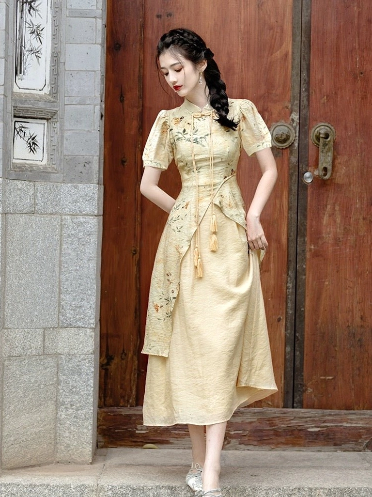 Three-quarter view of an apricot beige modern Hanfu midi dress featuring a stand collar, short puff sleeves, asymmetrical printed layer, tassel decoration, and flowing skirt