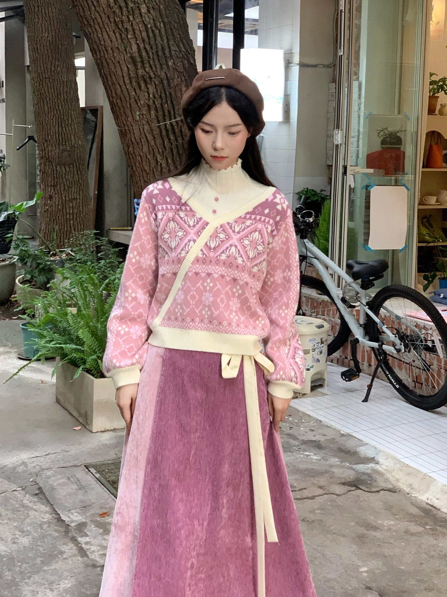 Front view of a pink modern Hanfu-inspired floral fair isle wrap knit sweater styled with a mauve skirt, beret, and outdoor garden accessories