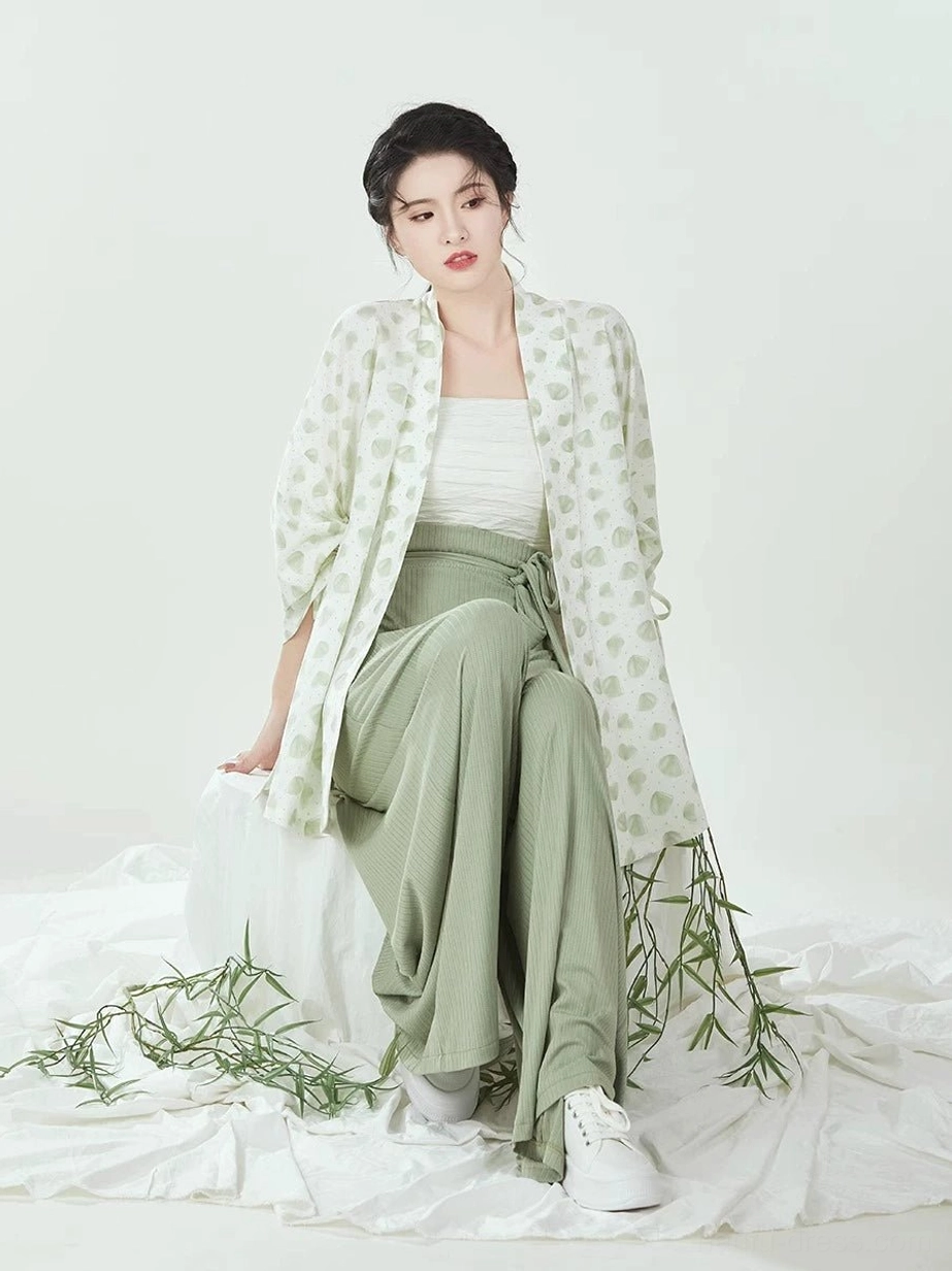 Seated model wearing complete modern Hanfu set with leaf-print winged sleeve top, camisole, sage green pants, and white sneakers