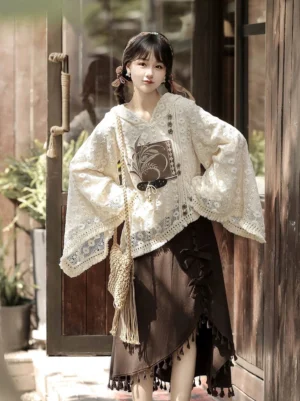 Front three-quarter view of a woman wearing a modern Hanfu two-piece outfit with beige floral lace hooded top, wheat grain detail, brown asymmetric tassel skirt, and side bow in a vintage courtyard