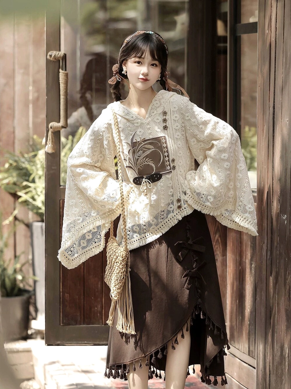 Front three-quarter view of a woman wearing a modern Hanfu two-piece outfit with beige floral lace hooded top, wheat grain detail, brown asymmetric tassel skirt, and side bow in a vintage courtyard