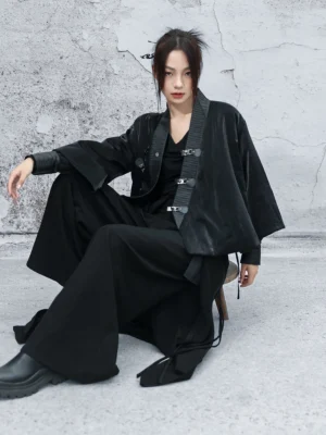 Modern Chinese-style hanfu wuxia cropped coat shown from the front while seated, featuring glossy black pleated fabric, an angled wrap collar, metal buckle closures, and wide bell sleeves styled with black trousers and boots