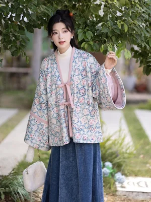 Front three-quarter view of a modern Jin-style Hanfu cardigan and dark blue velvet elastic waist skirt set worn in a garden, featuring pastel floral geometric print, pink ribbon ties, wide sleeves, and traditional hairpin styling