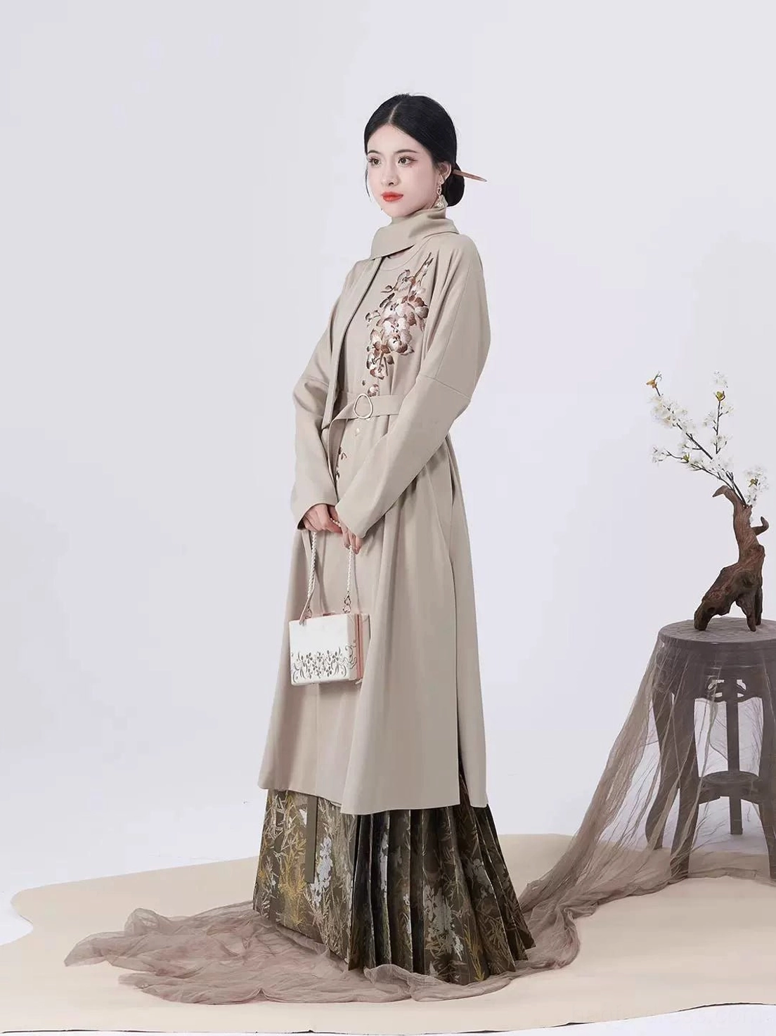 Full-body side view of modern Ming Dynasty Hanfu outfit with stand-collar taupe trench coat, high-waisted pleated mamian skirt, and mini handbag