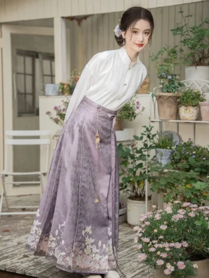Modern Ming Dynasty Hanfu set shown from a left rear three-quarter view with a dusty purple mamianqun horse face skirt, subtle jacquard texture, white floral hem details, and white cross-collar blouse in an outdoor courtyard