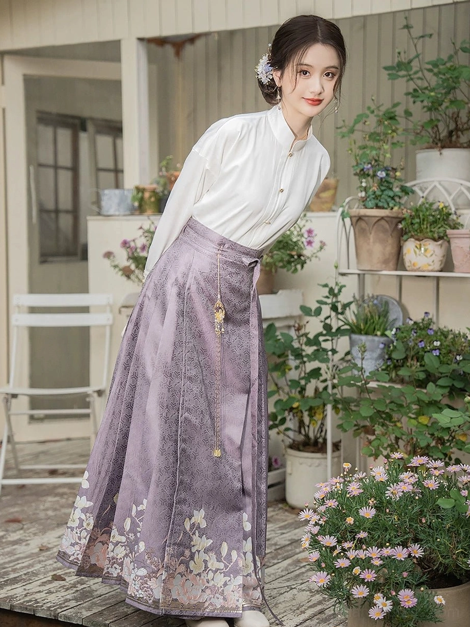 Modern Ming Dynasty Hanfu set shown from a left rear three-quarter view with a dusty purple mamianqun horse face skirt, subtle jacquard texture, white floral hem details, and white cross-collar blouse in an outdoor courtyard