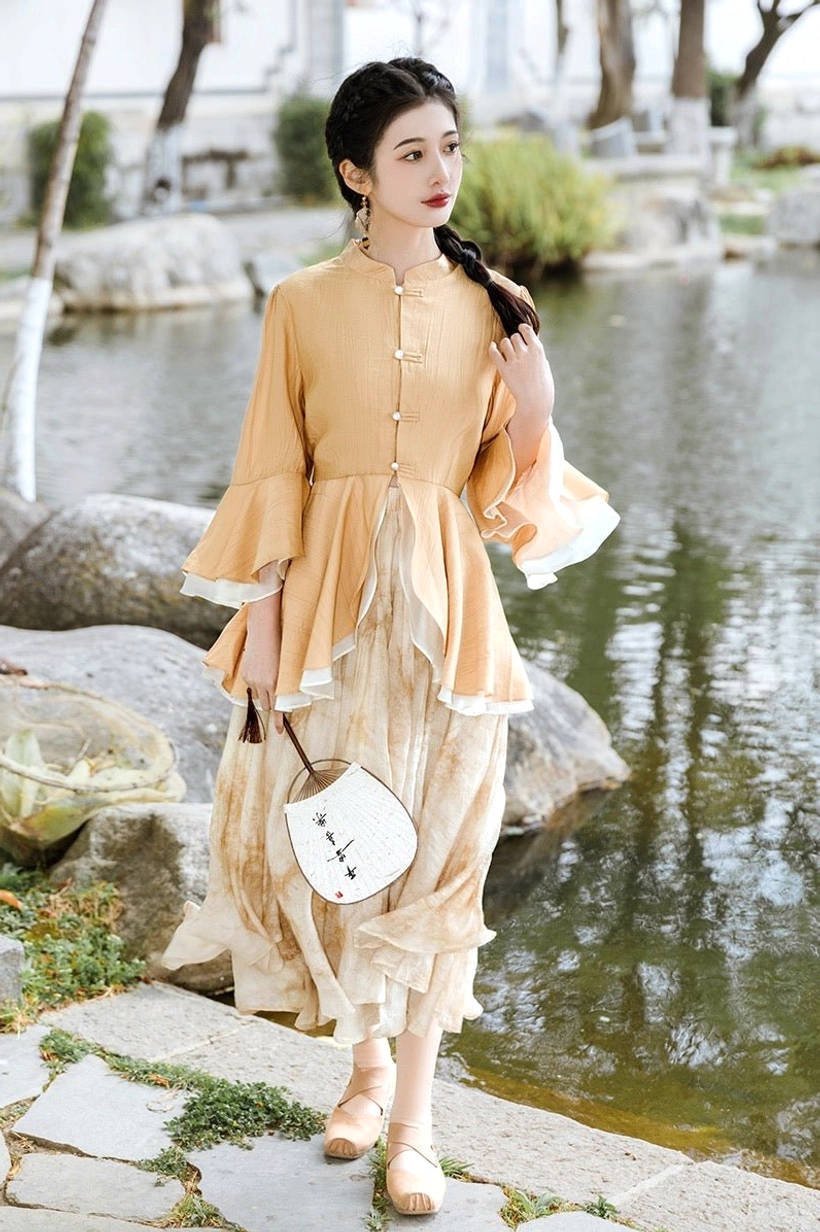 Full body modern Hanfu outfit featuring an apricot yellow Chinese stand collar top, flowing tie dye beige midi skirt, round Chinese fan, and beige flats