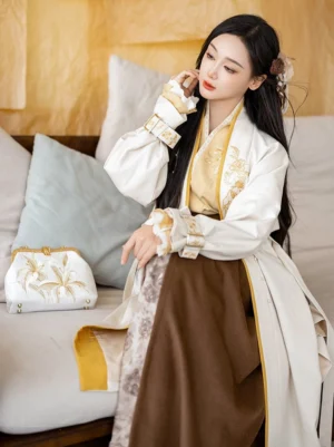 Modern Song Dynasty Hanfu outfit with cream winged sleeve jacket, mustard yellow trim, golden floral embroidery, and spiral wrap skirt shown on a seated model