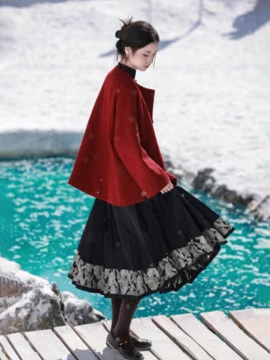 Back three-quarter view of a loose coral red Tang inspired hanfu jacket with flowing black pleated skirt and leather loafers