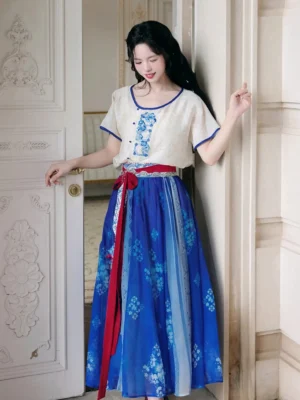 Modern Tang Hanfu two piece set with cream blue embroidered round neck top, royal blue floral long skirt, and red waist sash worn in a bright interior