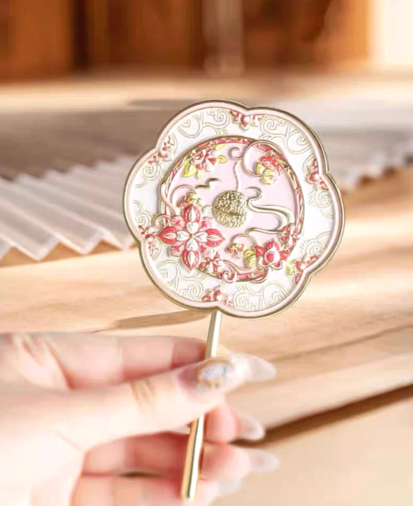 Hand holding a moonlit lotus Chinese hand mirror with auspicious cloud motifs and gold-toned handle beside a traditional folding fan