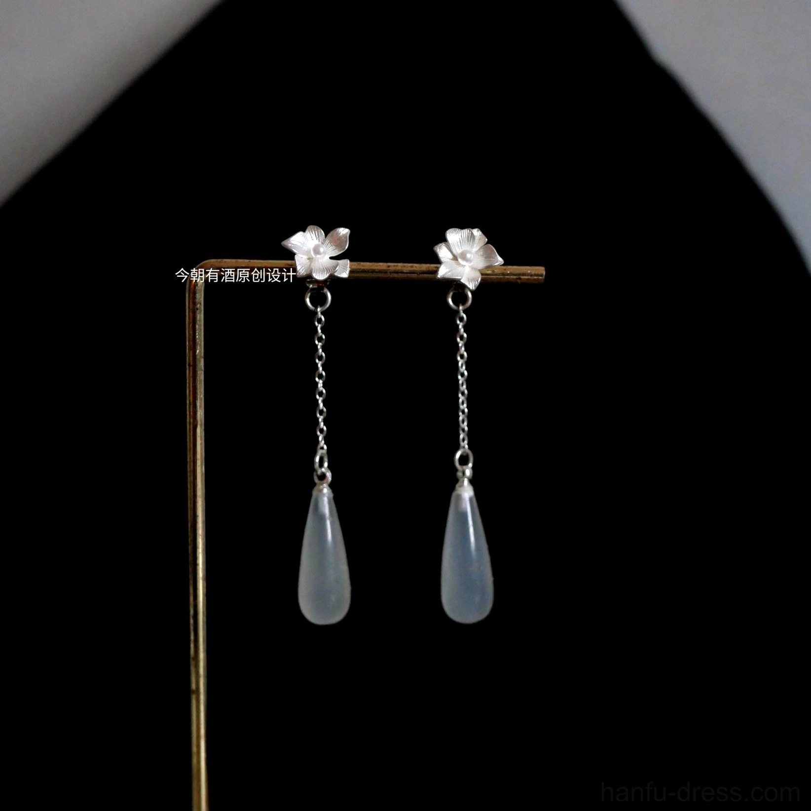 Floral Hanfu dangle earrings with silver flower studs, delicate chains, and opaque light blue teardrop pendants displayed on a gold jewelry stand against a black background