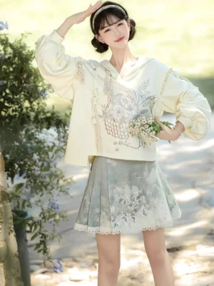 Three quarter view of cream embroidered Hanfu hoodie with tassels and green horse face skirt in a garden setting