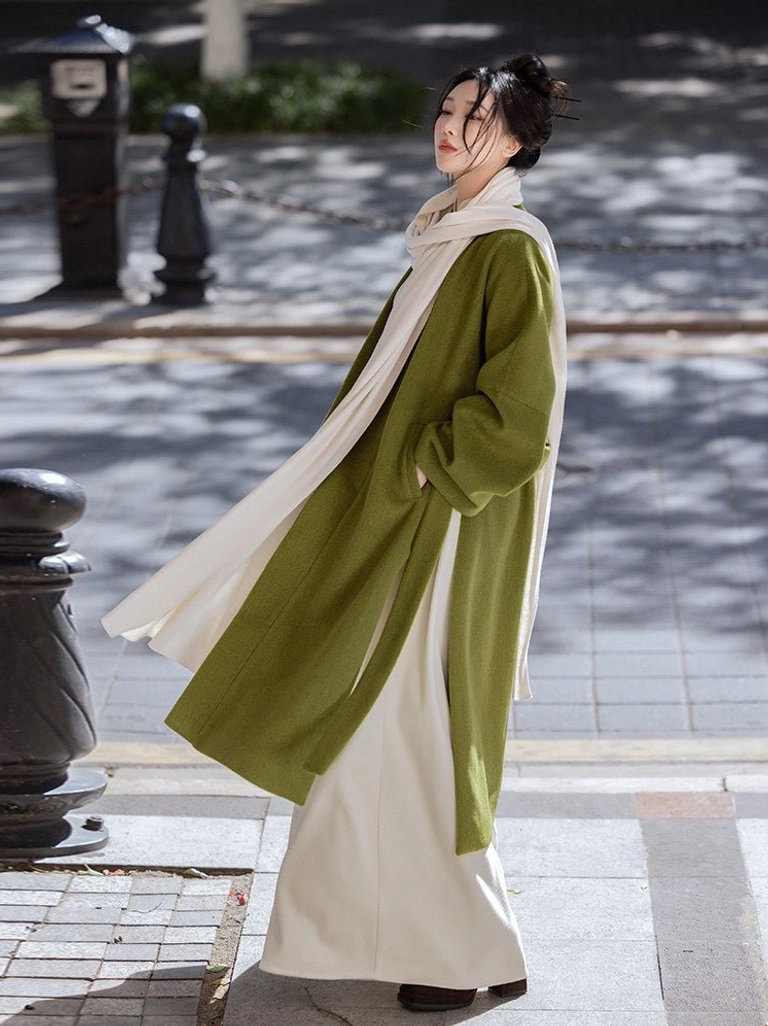 Side profile of a woman wearing a relaxed moss green modern Hanfu coat with flowing off-white scarf in an urban setting