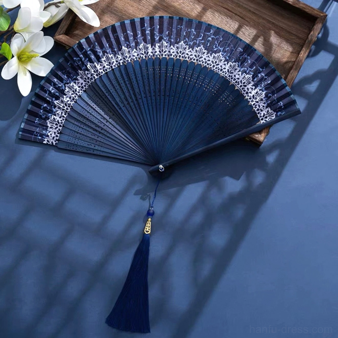 Navy blue Chinese folding hand fan with white lace-style floral trim, openwork bamboo ribs, blue tassel and gold charm displayed on a vintage wooden table
