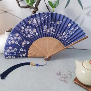 Navy sakura Chinese folding hand fan with white cherry blossom print, bamboo ribs, and blue tassel displayed on a traditional tea table