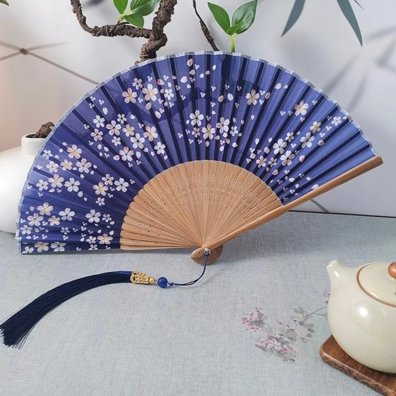 Navy sakura Chinese folding hand fan with white cherry blossom print, bamboo ribs, and blue tassel displayed on a traditional tea table