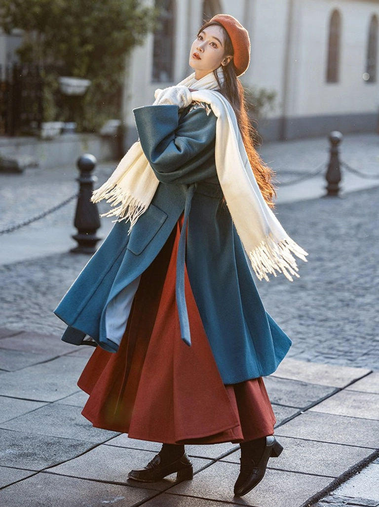 Side three-quarter view of a New Chinese Style winter outfit with a loose dark teal belted beizi coat, burgundy pleated wrap skirt, off-white scarf, red beret, and leather boots on a cobblestone street
