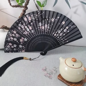 Black Chinese folding hand fan with pink and white cherry blossoms, bamboo ribs, carved details, long black tassel and metal pendant displayed beside a ceramic teapot and green plants